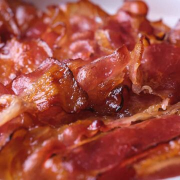 Close-up of several crispy, cooked strips of bacon overlapping on a white plate. The bacon appears shiny and slightly curled, with areas of golden brown and reddish color.
