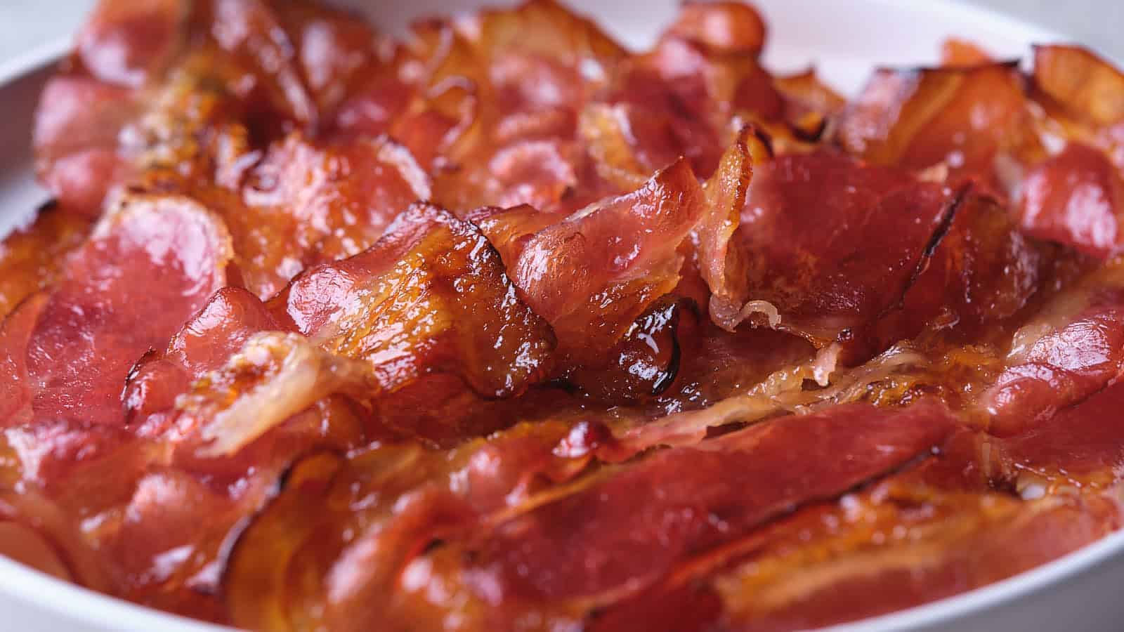 Close-up of several crispy, cooked strips of bacon overlapping on a white plate. The bacon appears shiny and slightly curled, with areas of golden brown and reddish color.