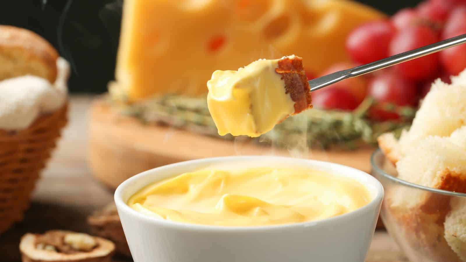 A close-up of a piece of bread dipped in a bowl of melted cheese, with bread, cheese, grapes, and herbs visible in the background. Steam is rising from the cheese.