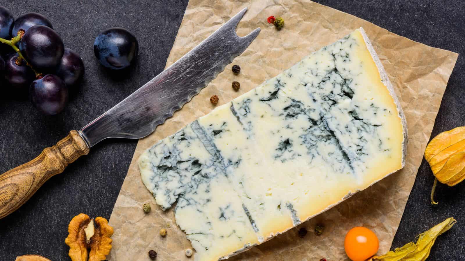 A wedge of blue cheese on parchment paper with a cheese knife, black grapes, walnuts, cape gooseberry, and scattered peppercorns on a dark surface.