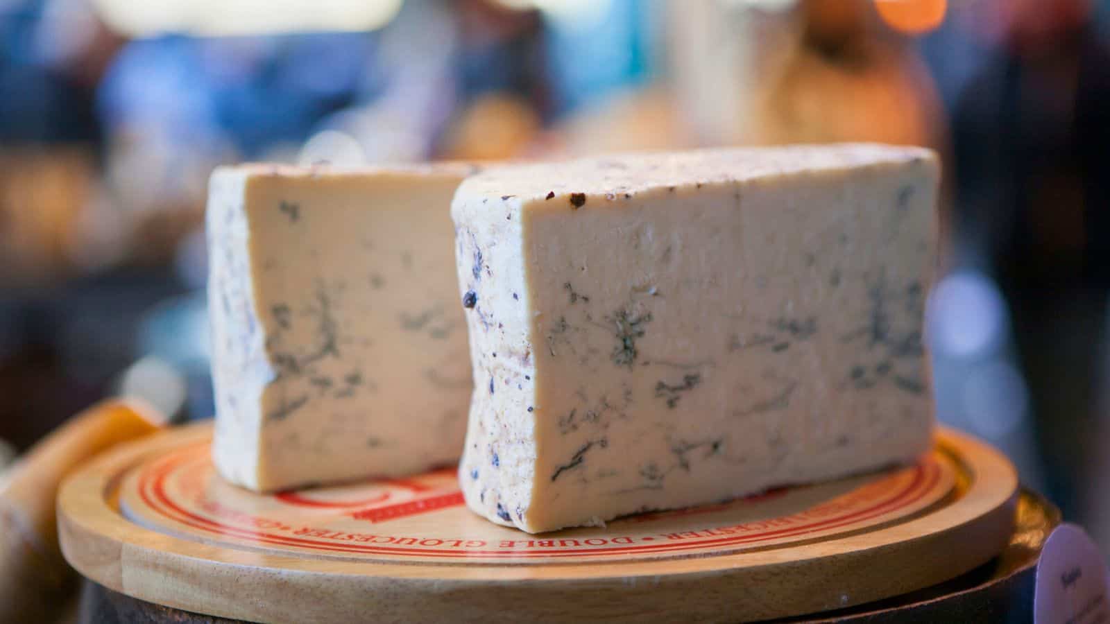 Two wedges of blue cheese with visible blue-green veins are displayed on a round wooden board with a branded paper liner. The background is blurred.
