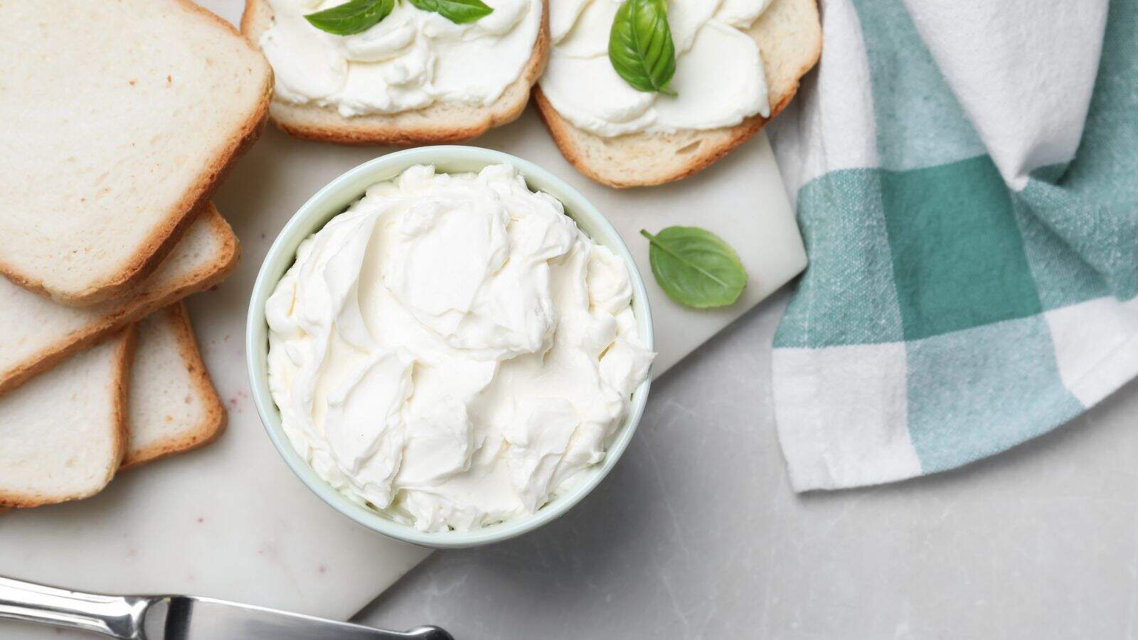 A bowl of cream cheese sits on a marble surface next to slices of white bread, some spread with cream cheese and basil leaves. A green and white checkered cloth and a butter knife are also visible.