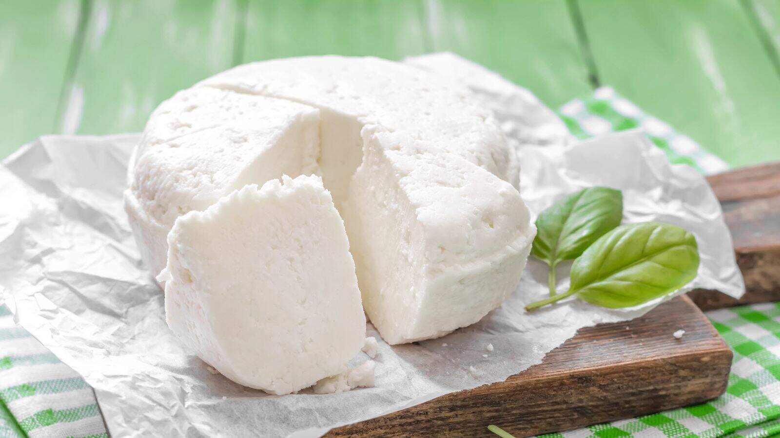 A round block of white cheese with a wedge cut out sits on crumpled parchment paper. Two green basil leaves are placed beside the cheese on a wooden board atop a green checkered cloth.
