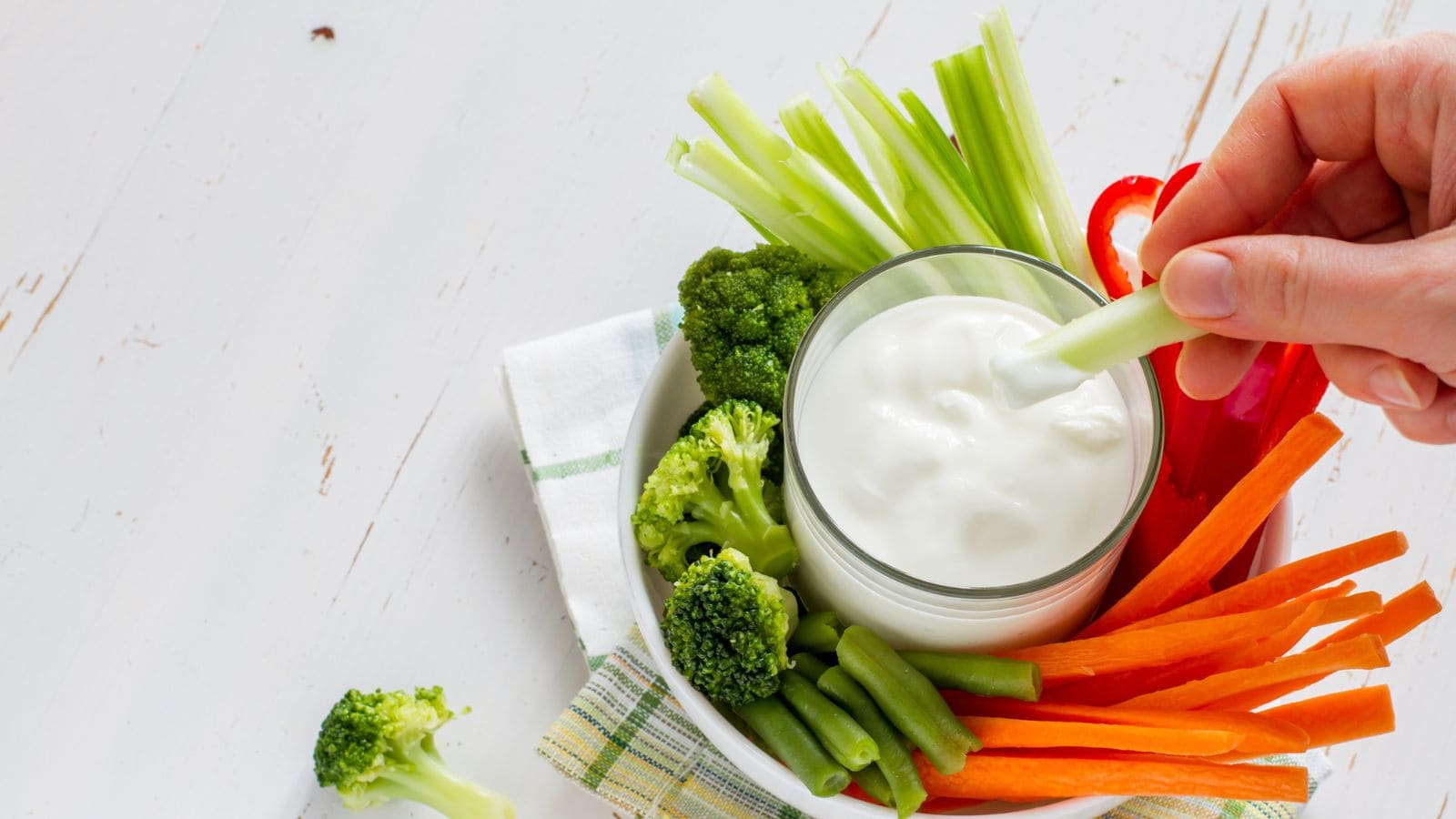A hand dips a celery stick into a glass of white dip, surrounded by broccoli, green beans, carrots, celery, and red pepper slices on a plate with a white and green napkin underneath.