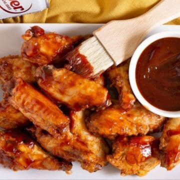 A white plate with glazed chicken wings sits on a yellow surface. A bowl of dark barbecue sauce and a basting brush coated in sauce rest beside the wings, with a can partially visible in the corner.