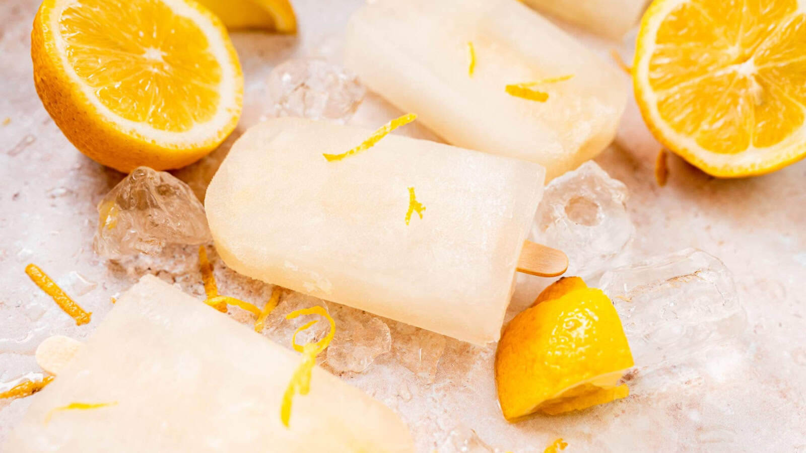 Several lemon popsicles rest on ice with lemon slices and zest scattered around them on a light surface.