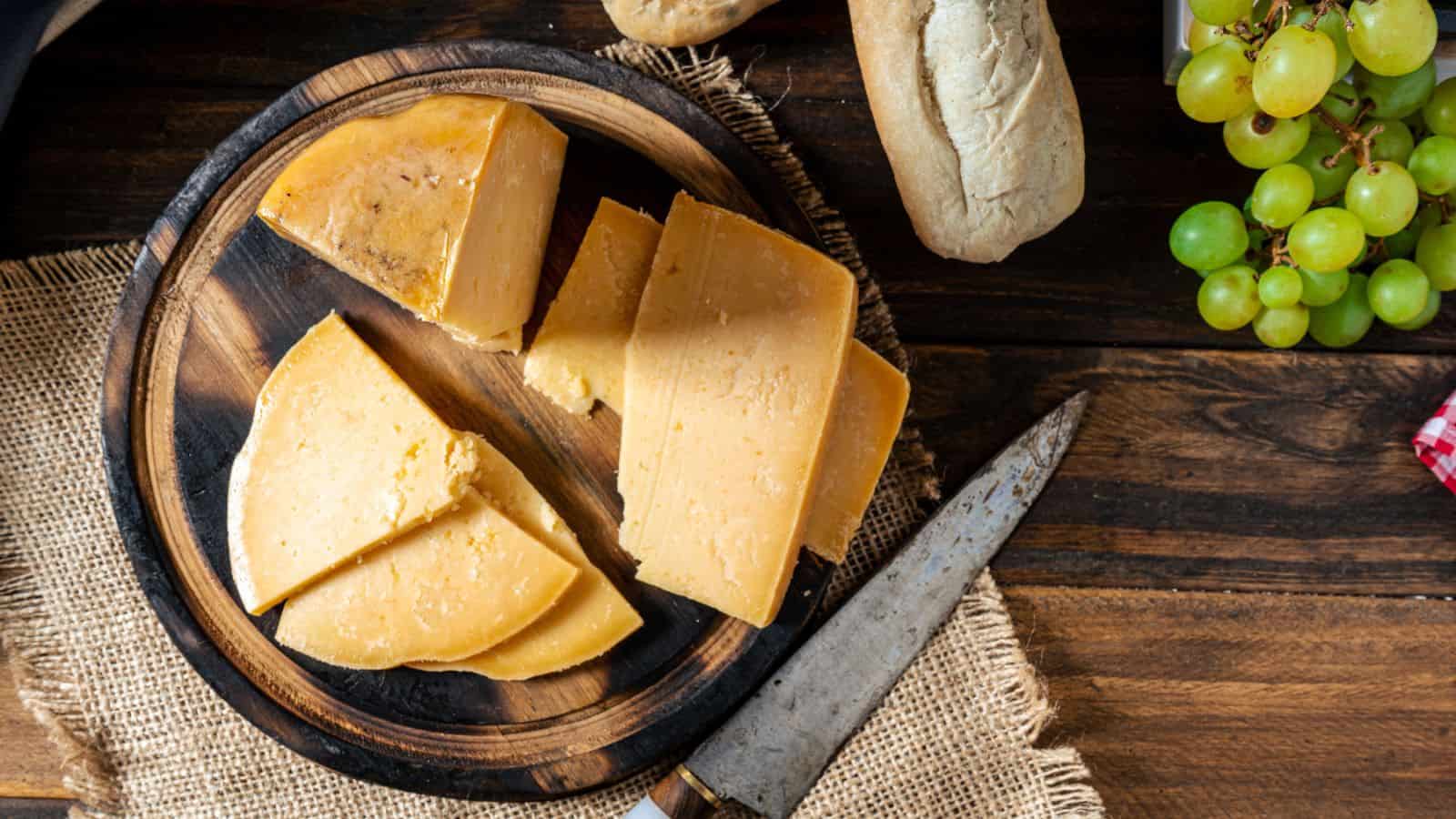 A round wooden board with several wedges of hard yellow cheese, a rustic knife, a couple of bread rolls, and a bunch of green grapes on a wooden table. The board sits on a piece of burlap.