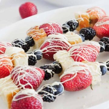 Fruit and cake skewers with strawberries, blueberries, blackberries, and cubes of cake are arranged on a white plate. The skewers are drizzled with white chocolate. A bowl of blackberries is in the background.