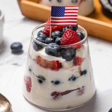 A glass filled with layers of yogurt, strawberries, and blueberries, topped with more berries and a small American flag. A spoon, a ramekin of blueberries, and a wooden tray are nearby on a marble surface.