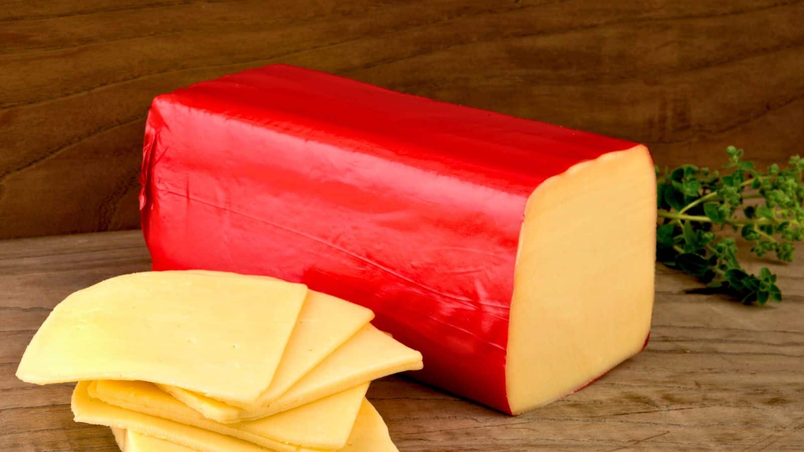 A rectangular block of yellow cheese with a red wax coating sits on a wooden surface. Several slices of the cheese are stacked in front of the block. A sprig of green herbs is in the background.