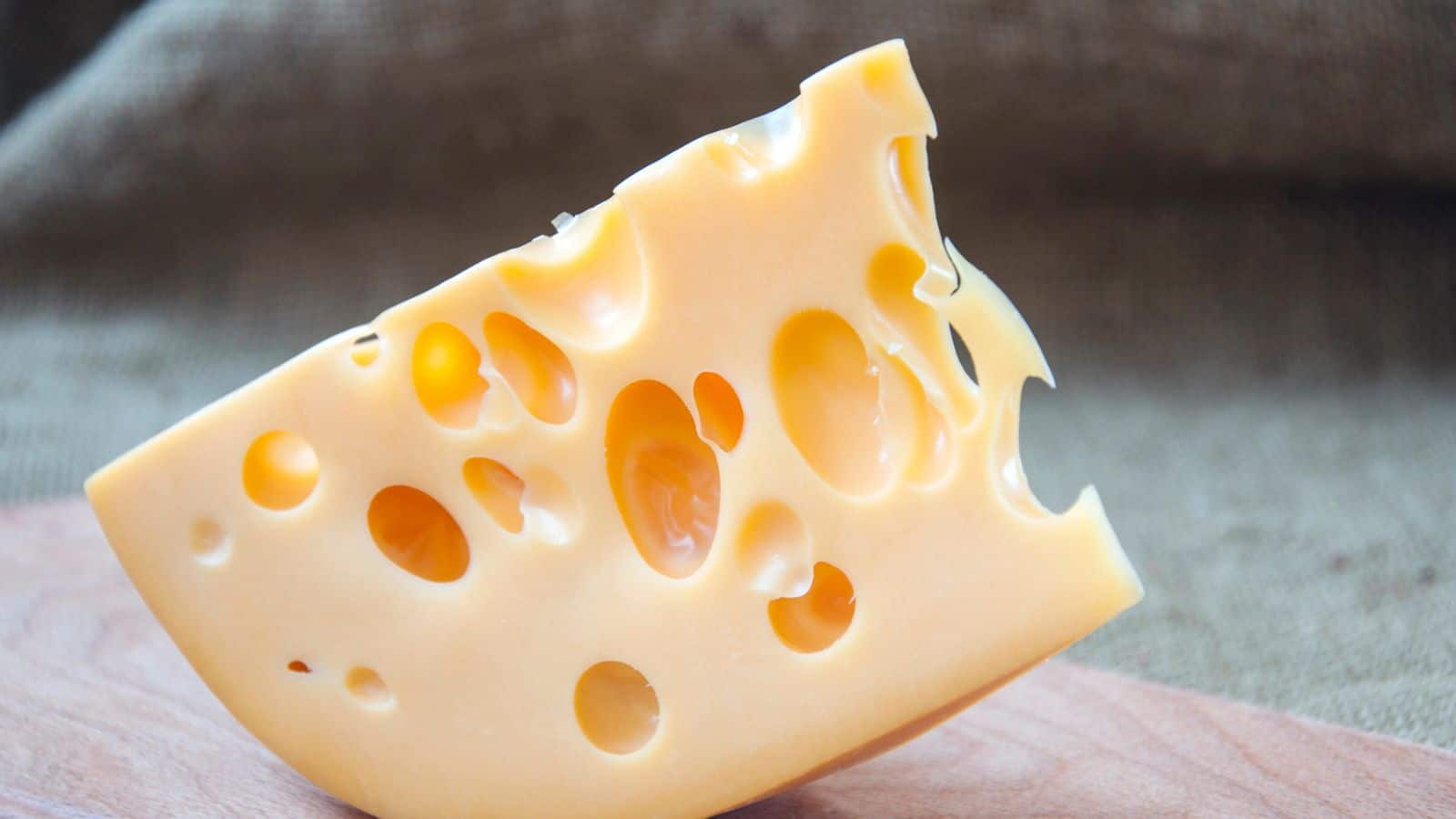 A wedge of Swiss cheese with multiple holes sits on a wooden surface, with a textured brown fabric background.