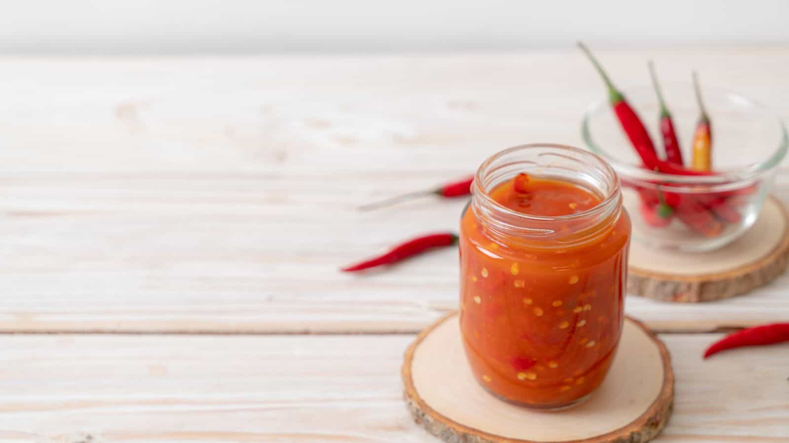 A glass jar filled with red chili sauce sits on a wooden coaster, with whole red chili peppers and another glass bowl in the background on a light wooden surface.