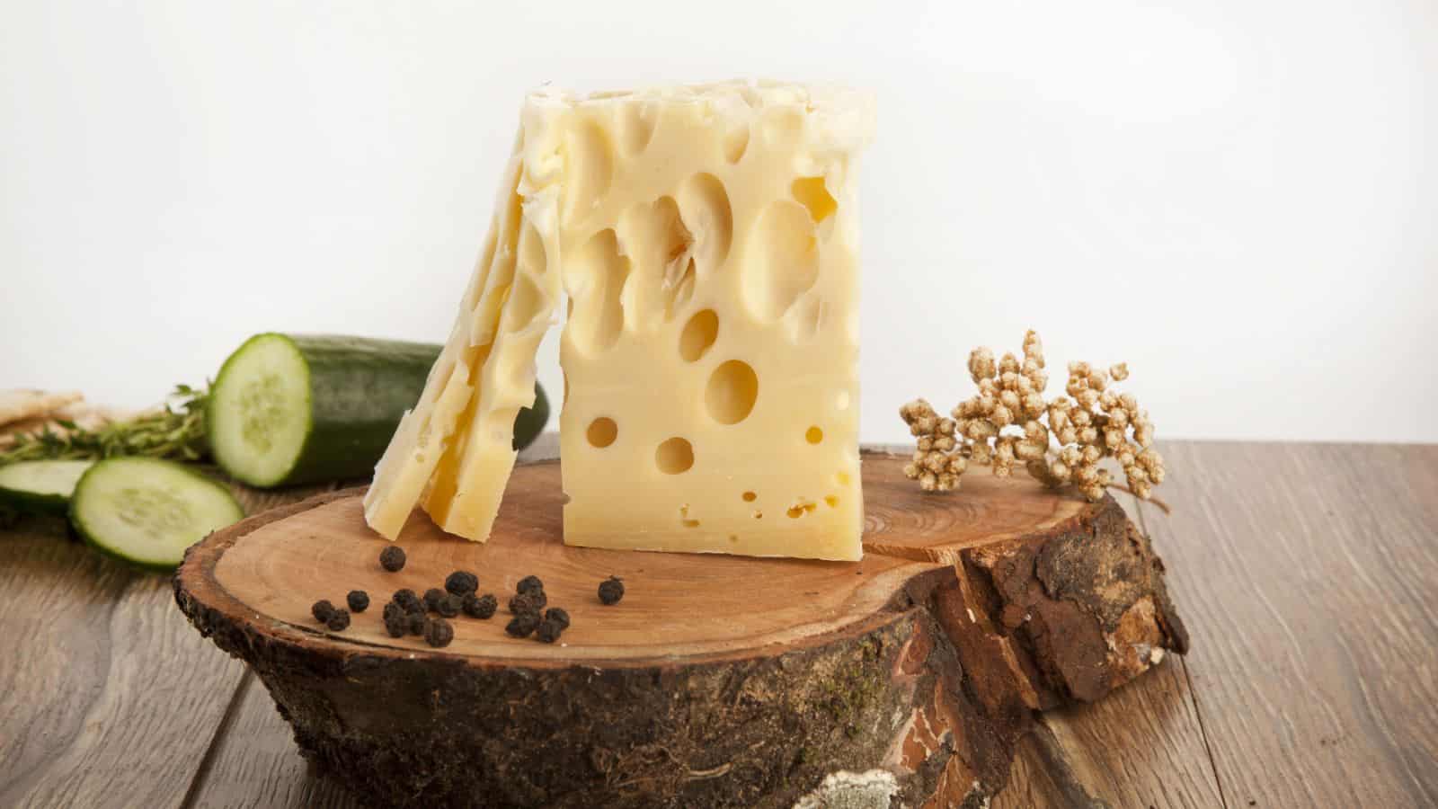A block and several slices of Swiss cheese with holes sit on a wooden board. Black peppercorns, cucumber slices, and a small cluster of white flowers are also on the board, with a plain white background.