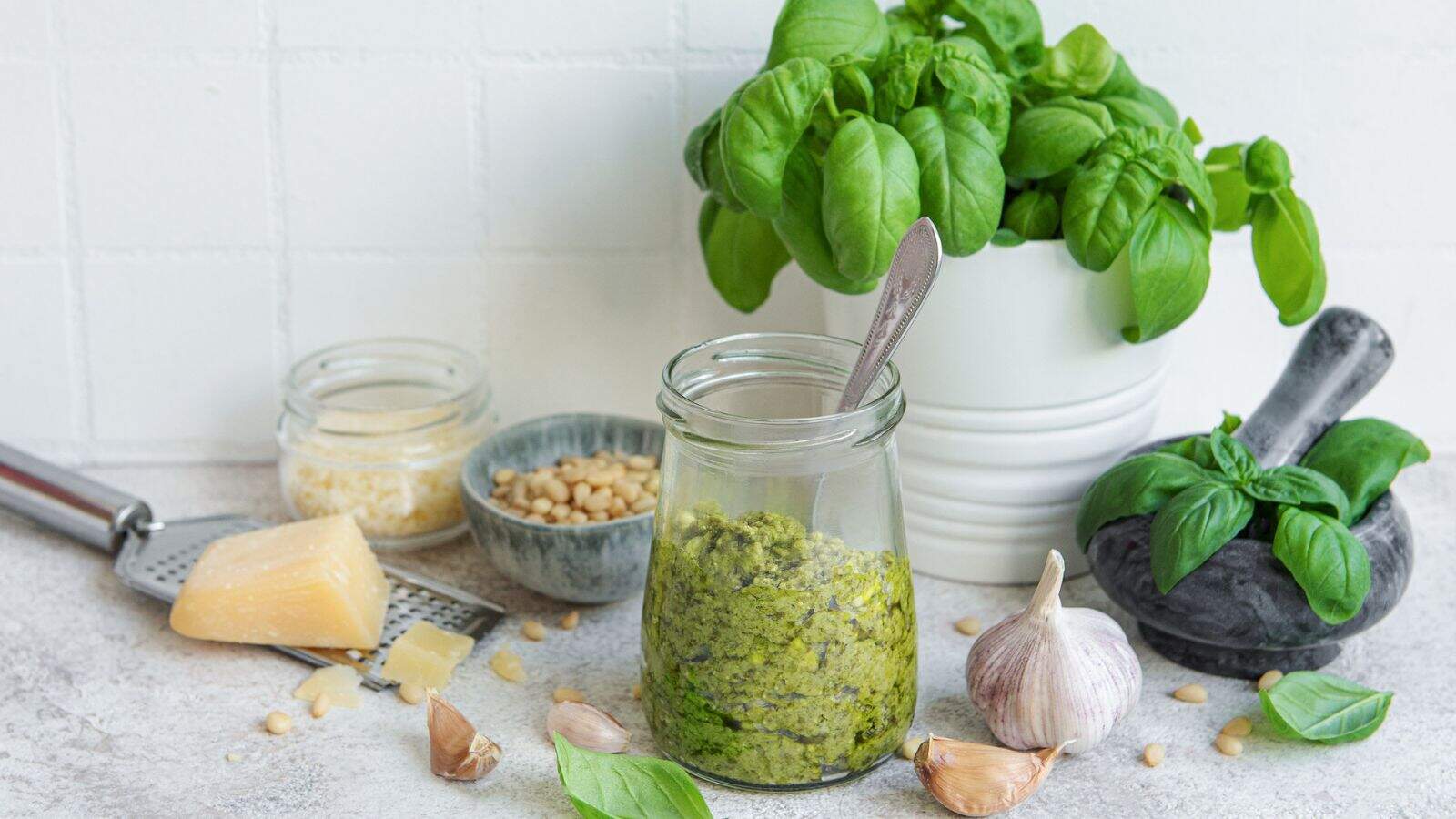 A jar of green pesto with a spoon inside is surrounded by fresh basil, garlic cloves, a wedge of cheese, grated cheese, pine nuts, and a mortar and pestle on a light kitchen surface.