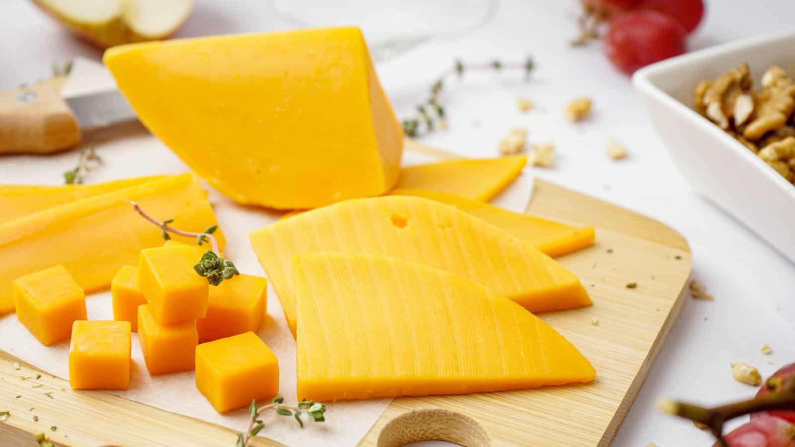 A wooden board with sliced, cubed, and wedge pieces of yellow cheddar cheese, garnished with herbs. Other foods, such as red grapes and walnuts, are visible in the background.