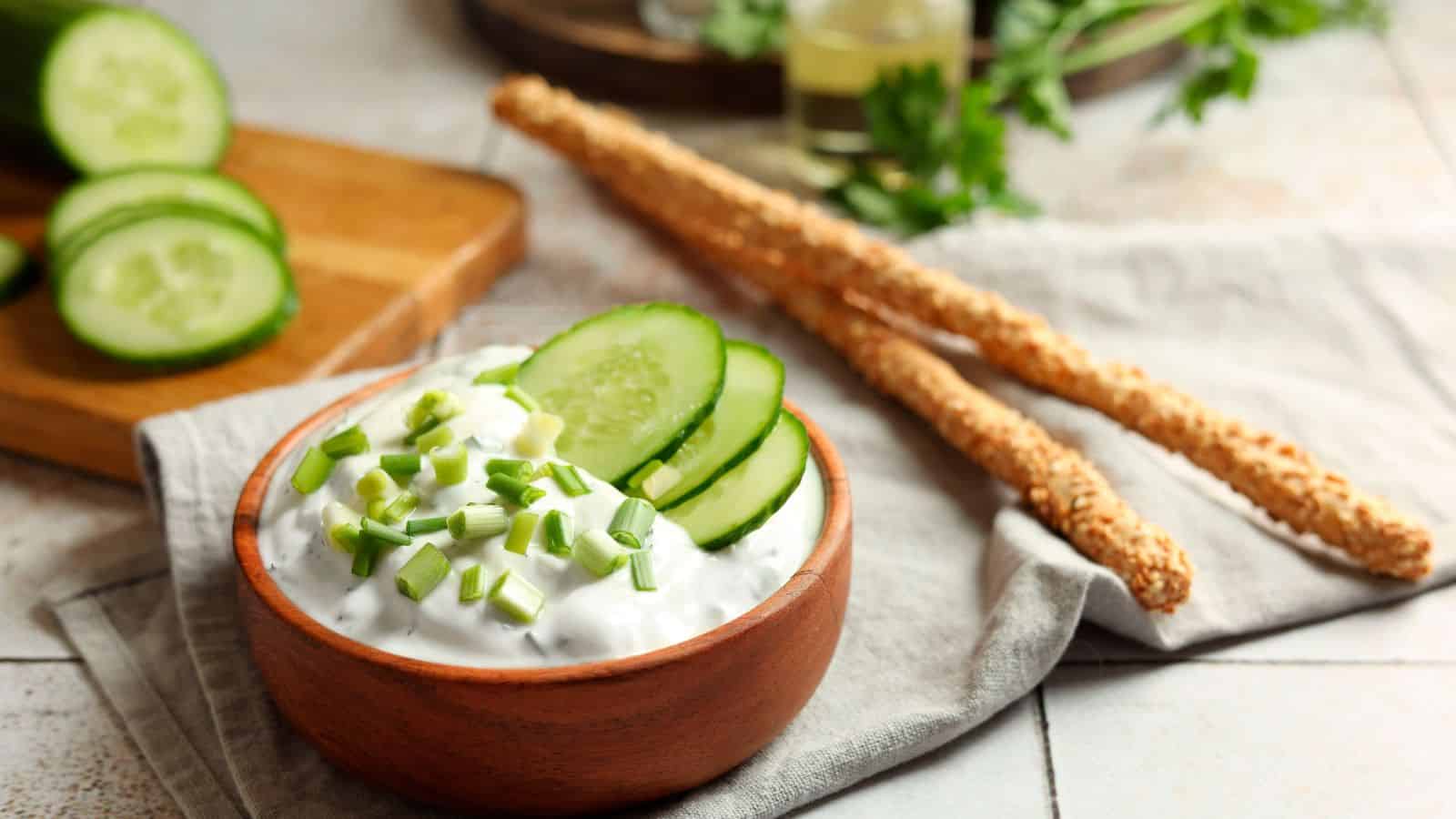 A wooden bowl filled with creamy dip topped with chopped chives and cucumber slices sits on a napkin. Two sesame breadsticks lie next to the bowl. Sliced cucumber and herbs are visible in the background.
