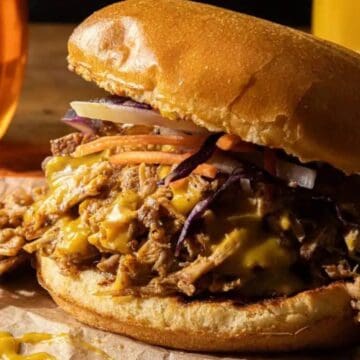 A pulled pork sandwich topped with shredded cheese and coleslaw sits on brown parchment paper. A glass of beer and a bottle of yellow sauce are partially visible in the background.
