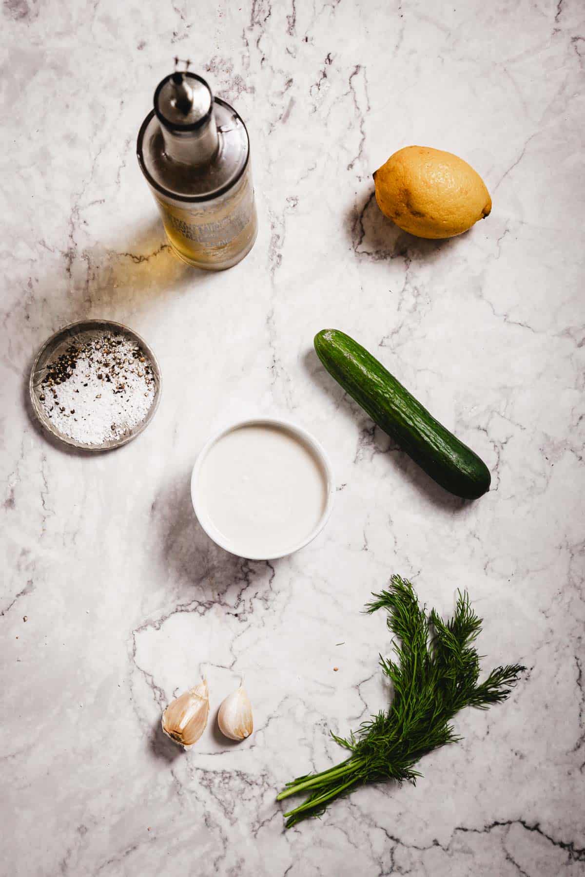 Ingredients for homemade Tzatziki Sauce on a marble countertop: a bottle of olive oil, a lemon, small cucumber, bowl of white sauce, dish of salt and pepper, two garlic cloves, and fresh dill.