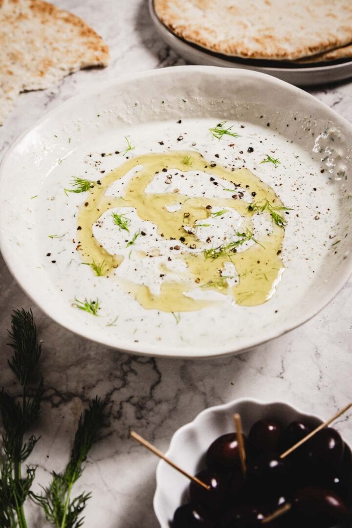 A white bowl filled with creamy Tzatziki Sauce, garnished with olive oil, dill, and cracked black pepper, sits on a marble surface. Pita bread, fresh dill, and a bowl of dark olives are arranged nearby.