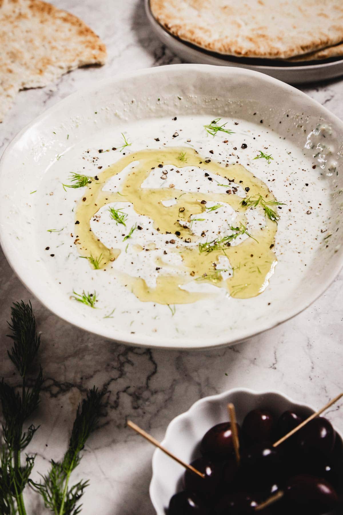 A white bowl filled with creamy Tzatziki Sauce, garnished with olive oil, dill, and cracked black pepper, sits on a marble surface. Pita bread, fresh dill, and a bowl of dark olives are arranged nearby.