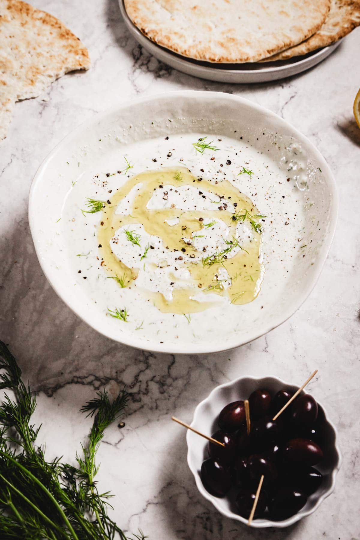 A white bowl of creamy Tzatziki Sauce, topped with olive oil, pepper, and dill, sits on a marble surface beside kalamata olives on toothpicks, fresh dill, and pieces of flatbread.