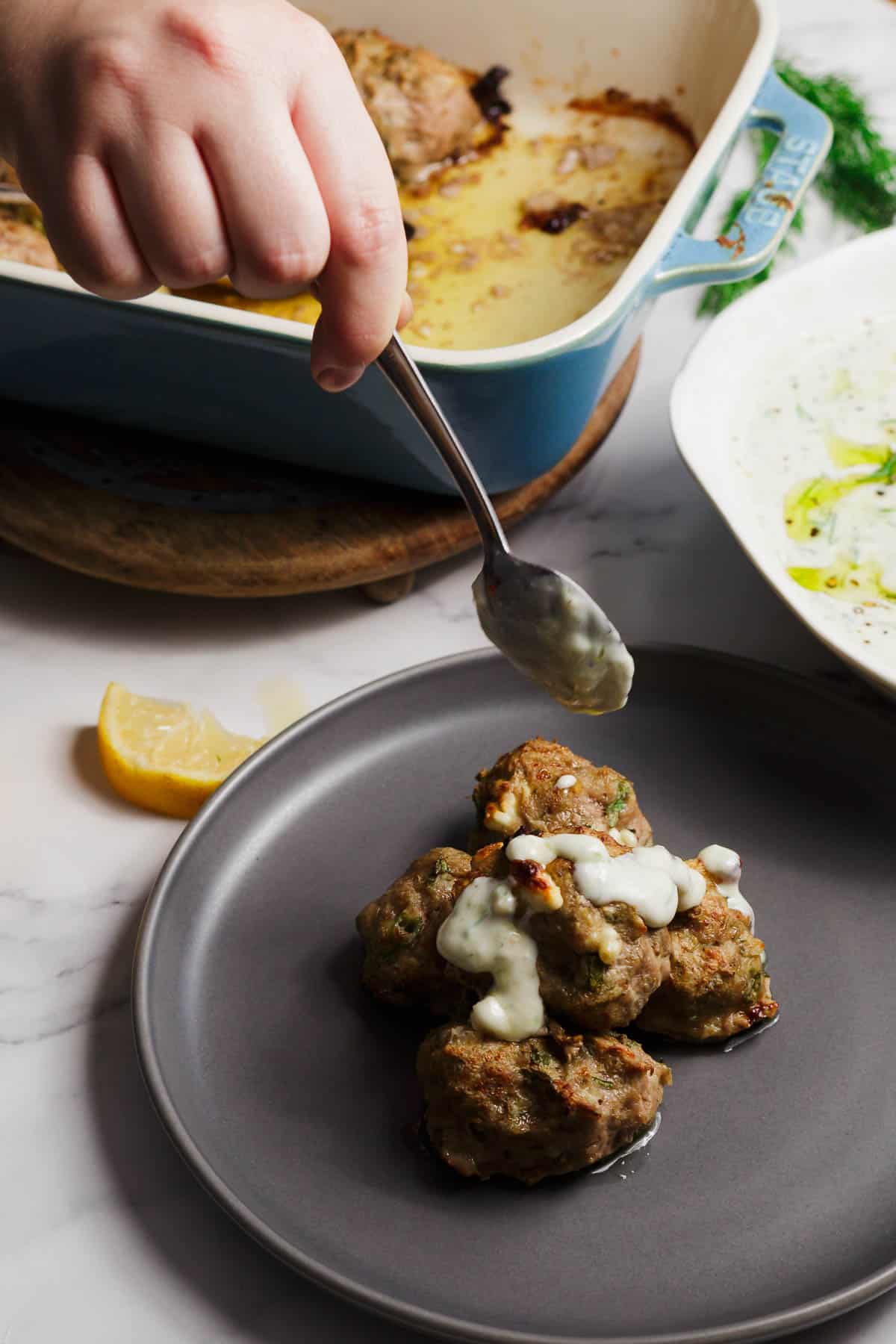 A hand uses a spoon to add creamy Tzatziki sauce to meatballs on a gray plate. In the background, a blue baking dish with more meatballs and a wedge of lemon rest on a marble surface.