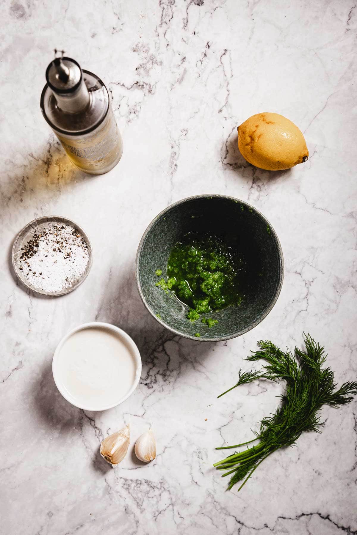 A marble surface displays a bottle of oil, a halved lemon, a bowl of green blended mixture, a dish of ground black pepper, tzatziki sauce in a white bowl, two garlic cloves, and a small bunch of fresh dill.