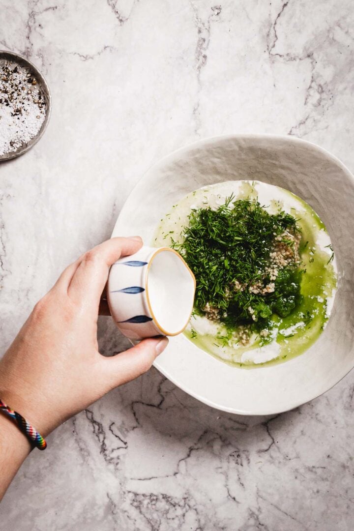 A hand pours liquid from a small pitcher into a white bowl containing chopped herbs, grated garlic, olive oil, and creamy ingredients on a marble countertop—classic components for making Tzatziki Sauce. A small dish of salt is also visible.