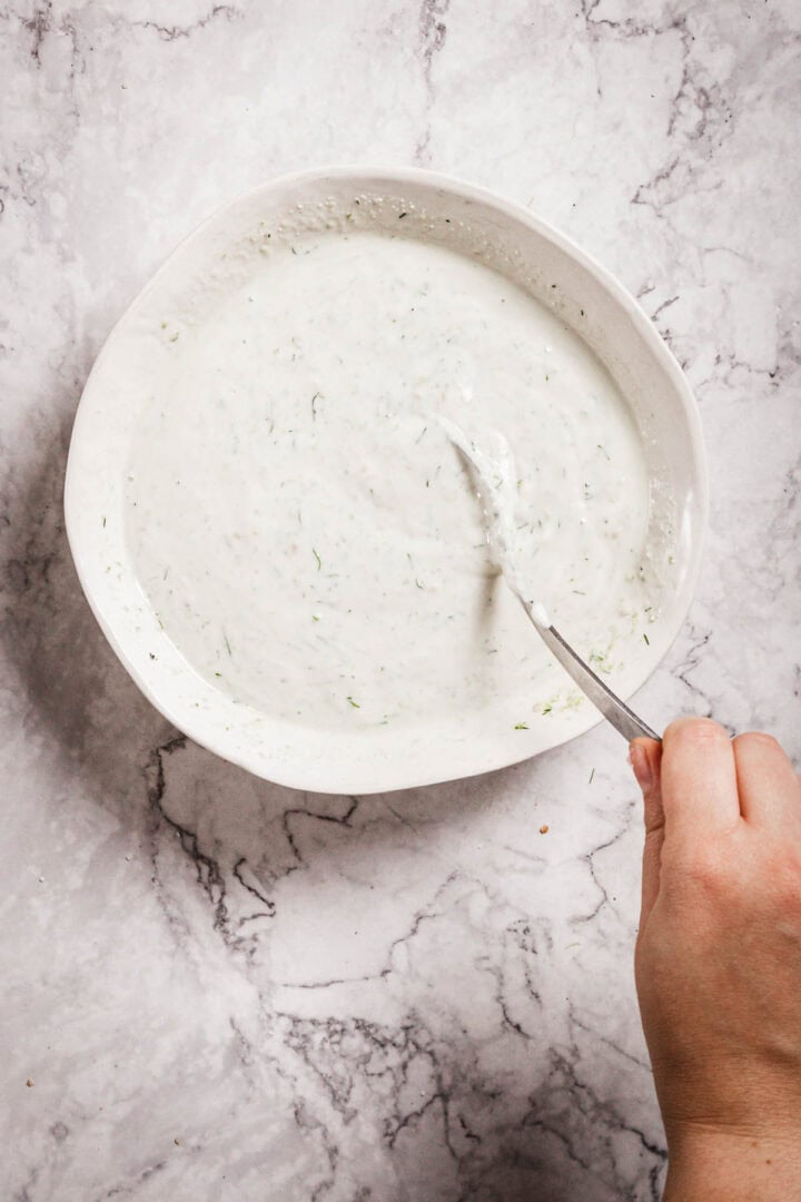 A hand holding a spoon is stirring a bowl of creamy Tzatziki Sauce with green herb flecks. The bowl sits on a light gray, marble-patterned surface.
