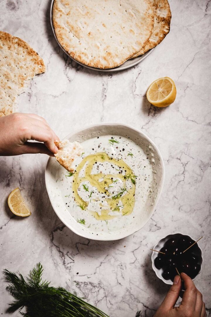 A hand dips flatbread into creamy Tzatziki Sauce garnished with olive oil and herbs; lemon wedges, olives, extra flatbread, and dill are artfully arranged on a marble surface nearby.