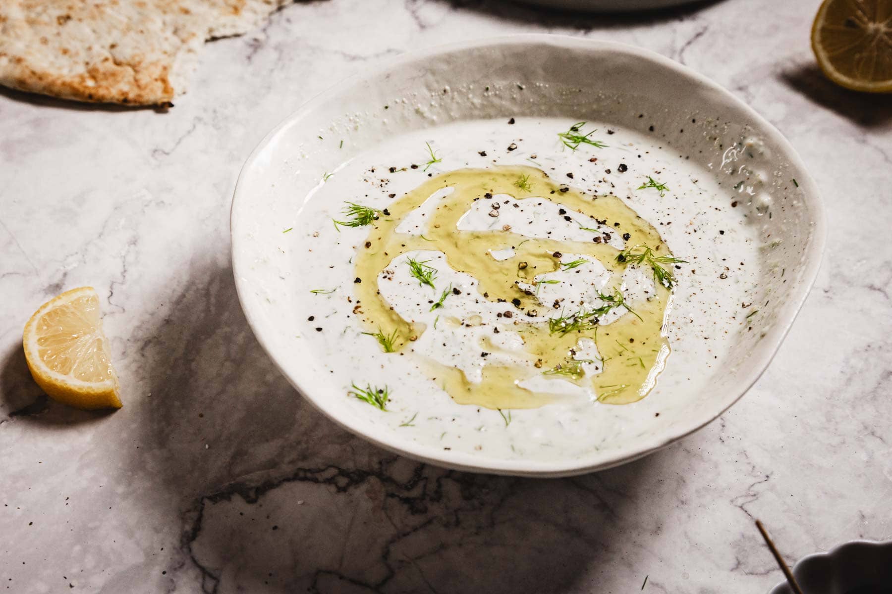 A white bowl filled with creamy Tzatziki Sauce, garnished with olive oil, dill, and black pepper, sits on a marble surface alongside lemon wedges and a piece of pita bread.