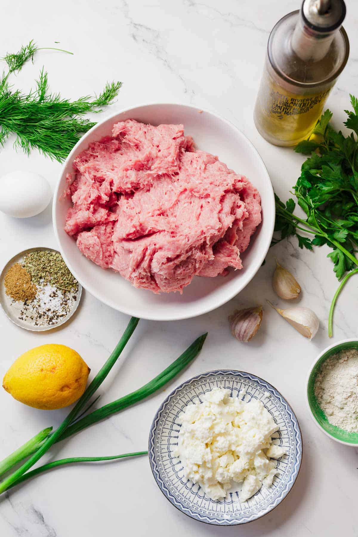A bowl of raw ground turkey surrounded by fresh herbs, a whole egg, a lemon, two garlic cloves, a bowl of breadcrumbs, a bowl of crumbled feta cheese, a bottle of olive oil, and assorted spices on a marble surface.