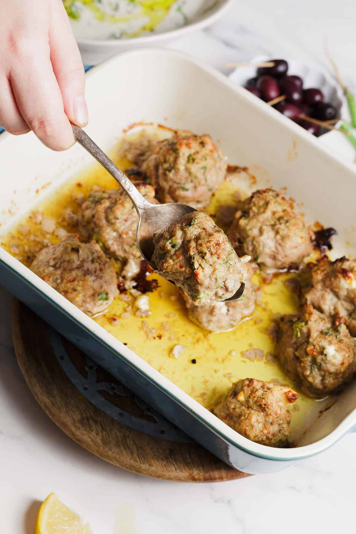 A hand uses a spoon to lift a baked meatball from a white rectangular dish containing several meatballs. The dish sits on a wooden trivet, with olives and part of a lemon visible nearby.