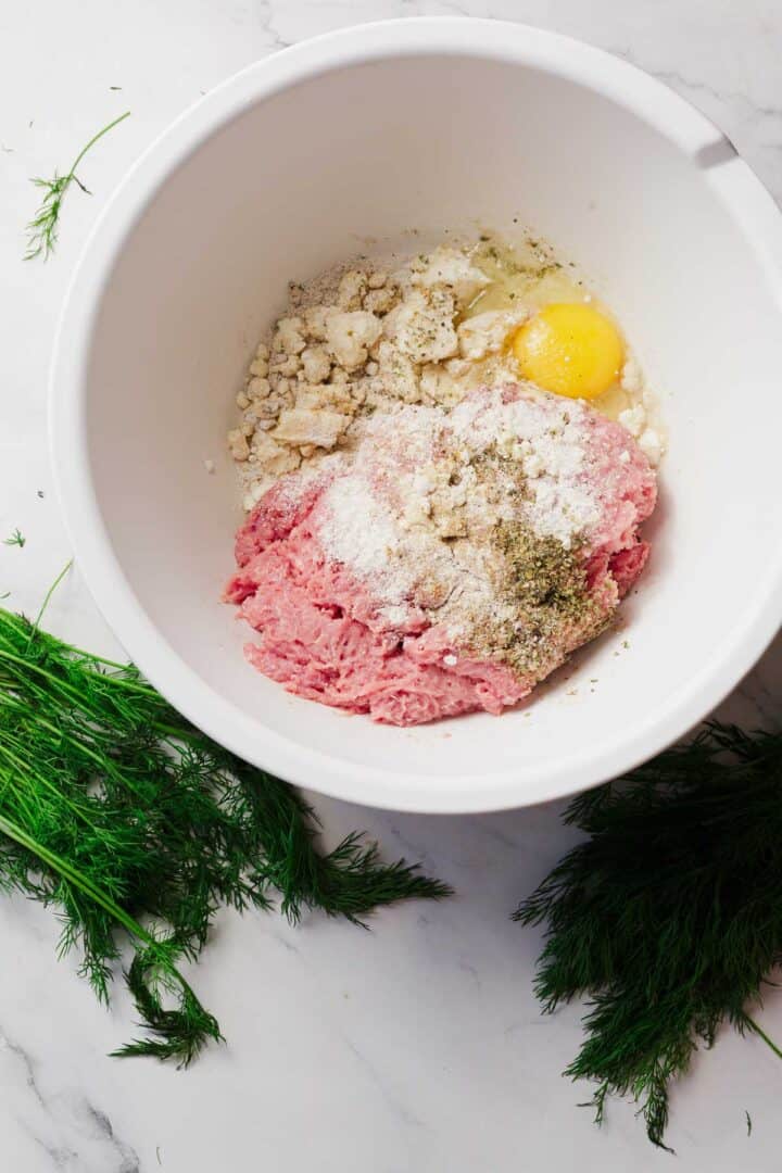 A white mixing bowl containing ground meat, an egg, bread crumbs, and seasonings sits on a marble surface with fresh dill sprigs nearby.
