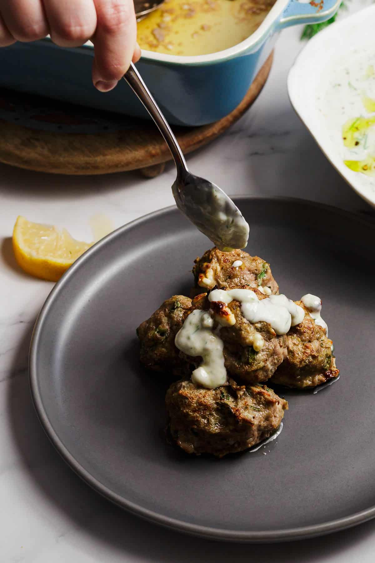 A hand holds a spoon, drizzling white sauce over several cooked meatballs piled on a gray plate. A lemon wedge and a blue baking dish are visible in the background.