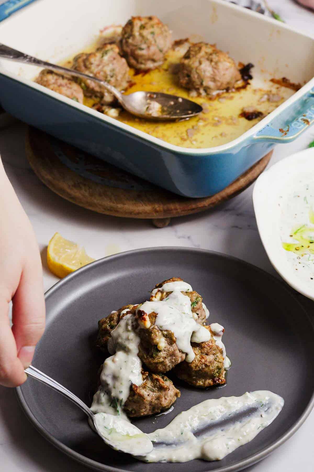 A person uses a fork to serve meatballs topped with creamy white sauce onto a gray plate. In the background, a blue baking dish with more meatballs and a serving spoon is visible. A lemon wedge is on the table.