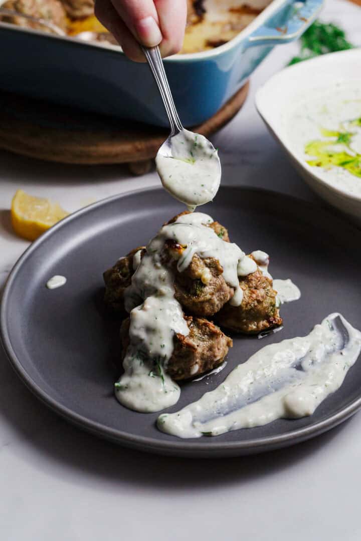 A hand holds a spoon, drizzling creamy white sauce over a stack of meatballs on a gray plate, with extra sauce spread on the side. A casserole dish and a lemon wedge are visible in the background.