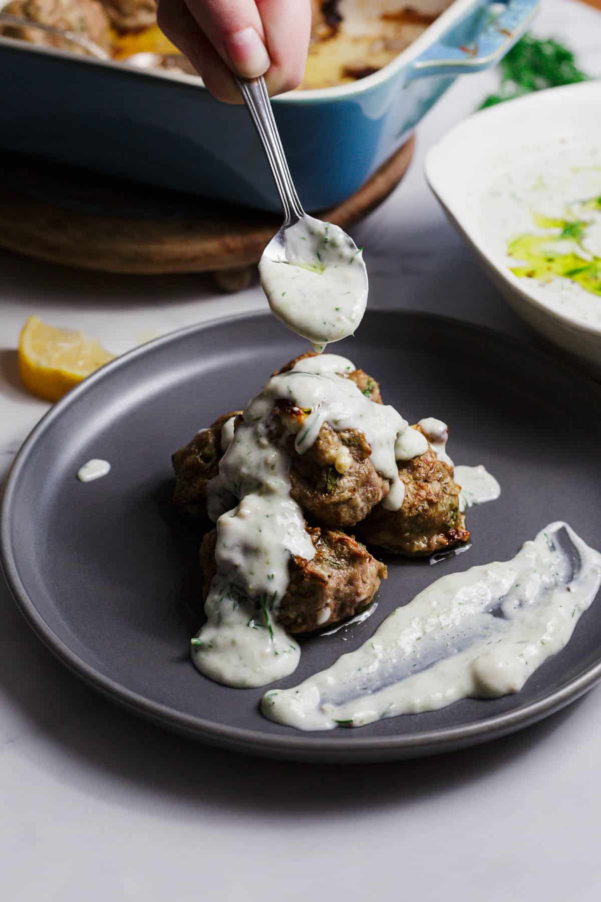 A hand holds a spoon, drizzling creamy white sauce over a stack of meatballs on a gray plate, with extra sauce spread on the side. A casserole dish and a lemon wedge are visible in the background.