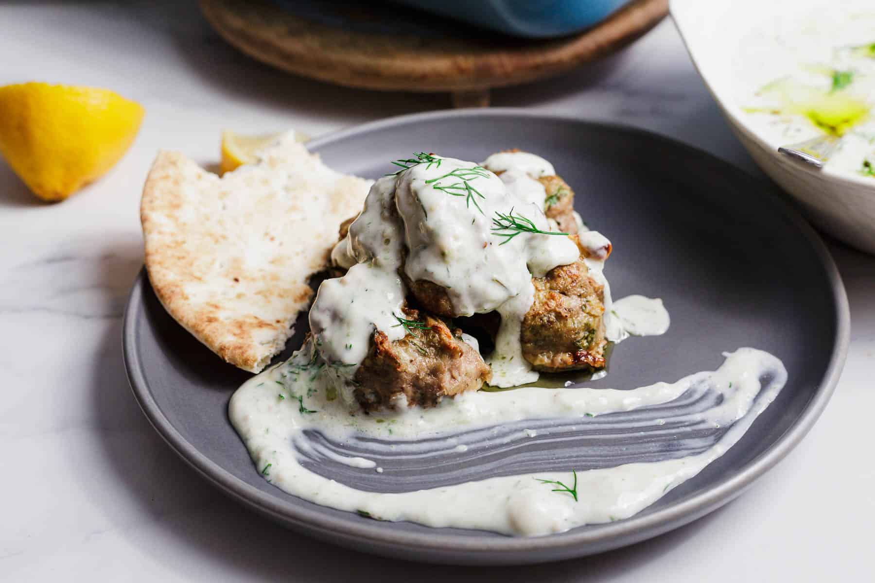 A gray plate with pita bread, grilled meat pieces topped with white tzatziki sauce, fresh dill, and a smear of sauce on the side. A lemon wedge and part of a salad bowl are visible in the background.