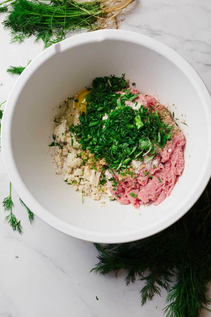 A white bowl contains ground meat, chopped herbs, breadcrumbs, egg, and seasonings, ready to be mixed. Fresh dill and herbs are scattered on a white marble surface nearby.