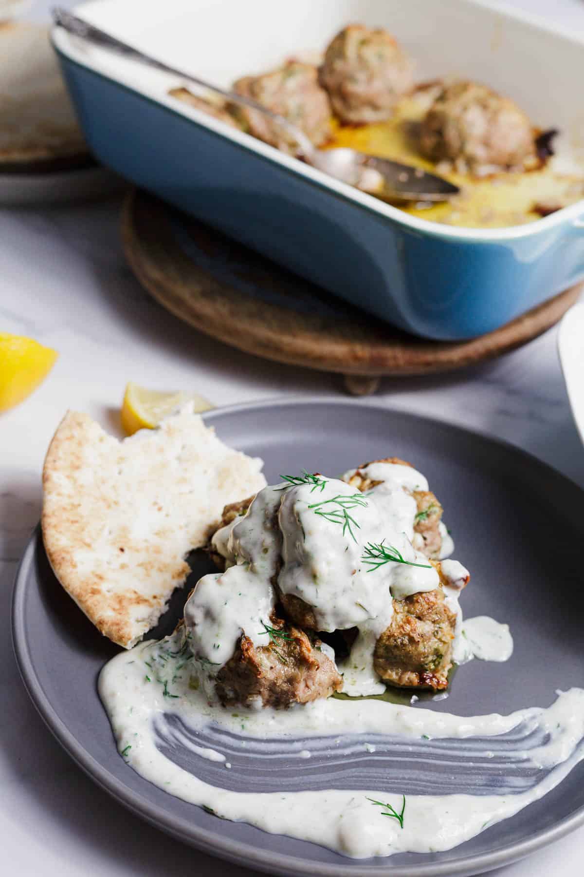 A gray plate with meatballs covered in white sauce, garnished with dill, and served with a piece of pita bread. In the background, a blue baking dish with more meatballs is visible.