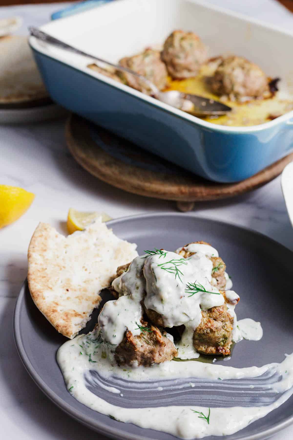A gray plate holds meatballs topped with creamy white sauce and fresh dill, served with pita bread. In the background, a blue baking dish with more meatballs is visible on a wooden trivet. A lemon wedge rests nearby.