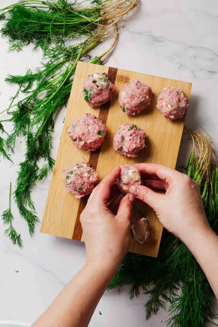 Two hands form raw meatballs on a wooden cutting board, with six meatballs arranged in rows. Fresh dill herbs and their roots are scattered on a white marble surface around the board.