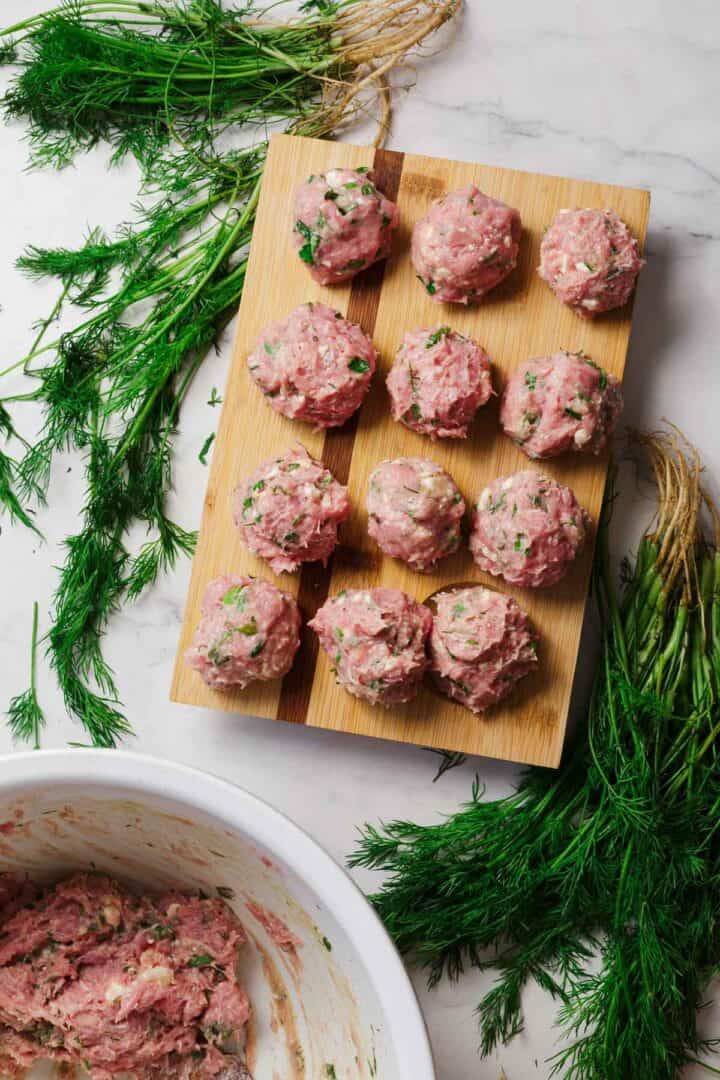 Raw meatballs with herbs are arranged on a wooden board. Fresh dill is scattered around the board on a marble surface. A white bowl with leftover meat mixture is visible in the lower left corner.