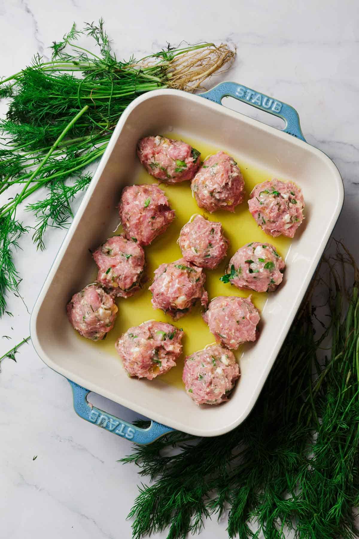 A blue Staub baking dish holds eleven raw meatballs in olive oil on a white marble surface, surrounded by fresh dill and other herbs.