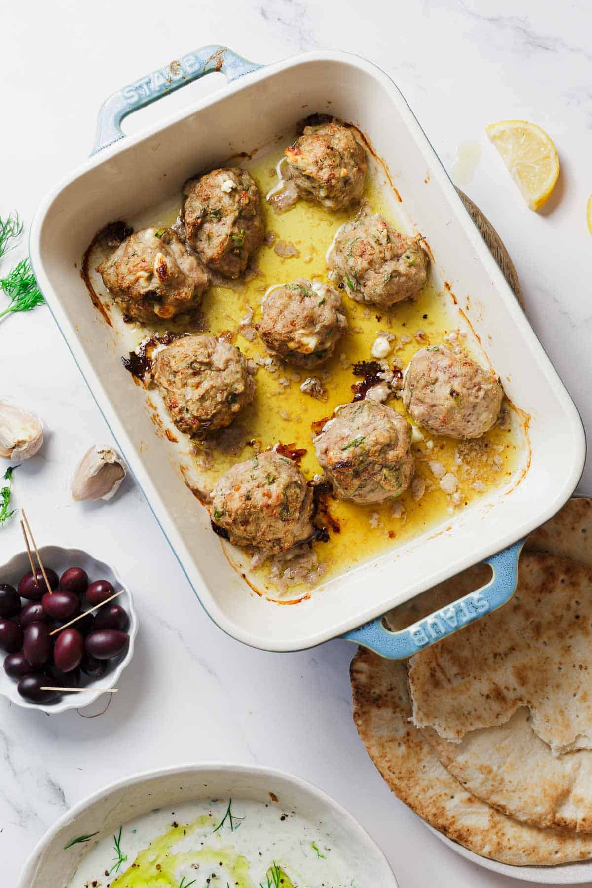A baking dish with nine cooked meatballs in oil and herbs, surrounded by pita bread, a bowl of olives, a bowl of yogurt dip, lemon wedge, garlic cloves, and fresh dill on a white marble surface.