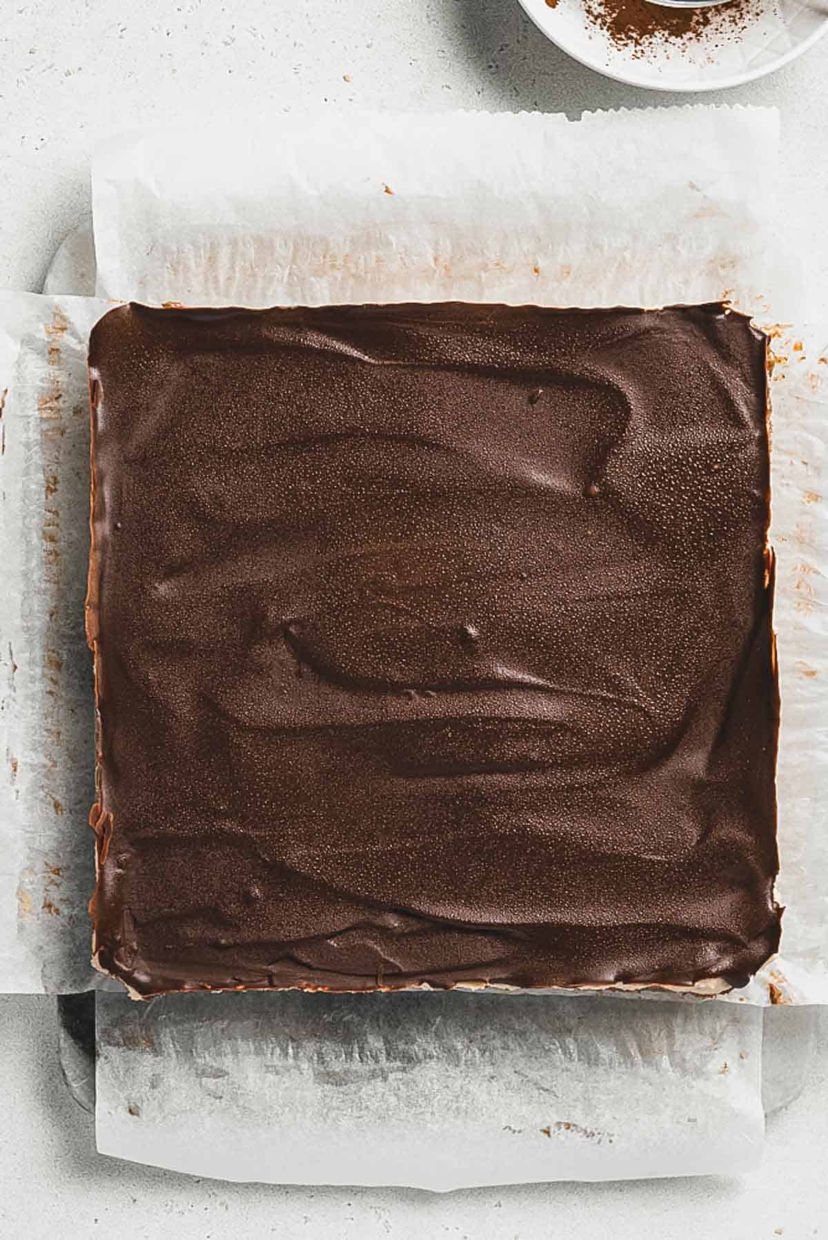 A square pan lined with parchment paper holds Tiramisu Protein Bars topped with a smooth layer of glossy chocolate. The surface appears even with visible swirl patterns. Part of a white plate is visible in the corner.