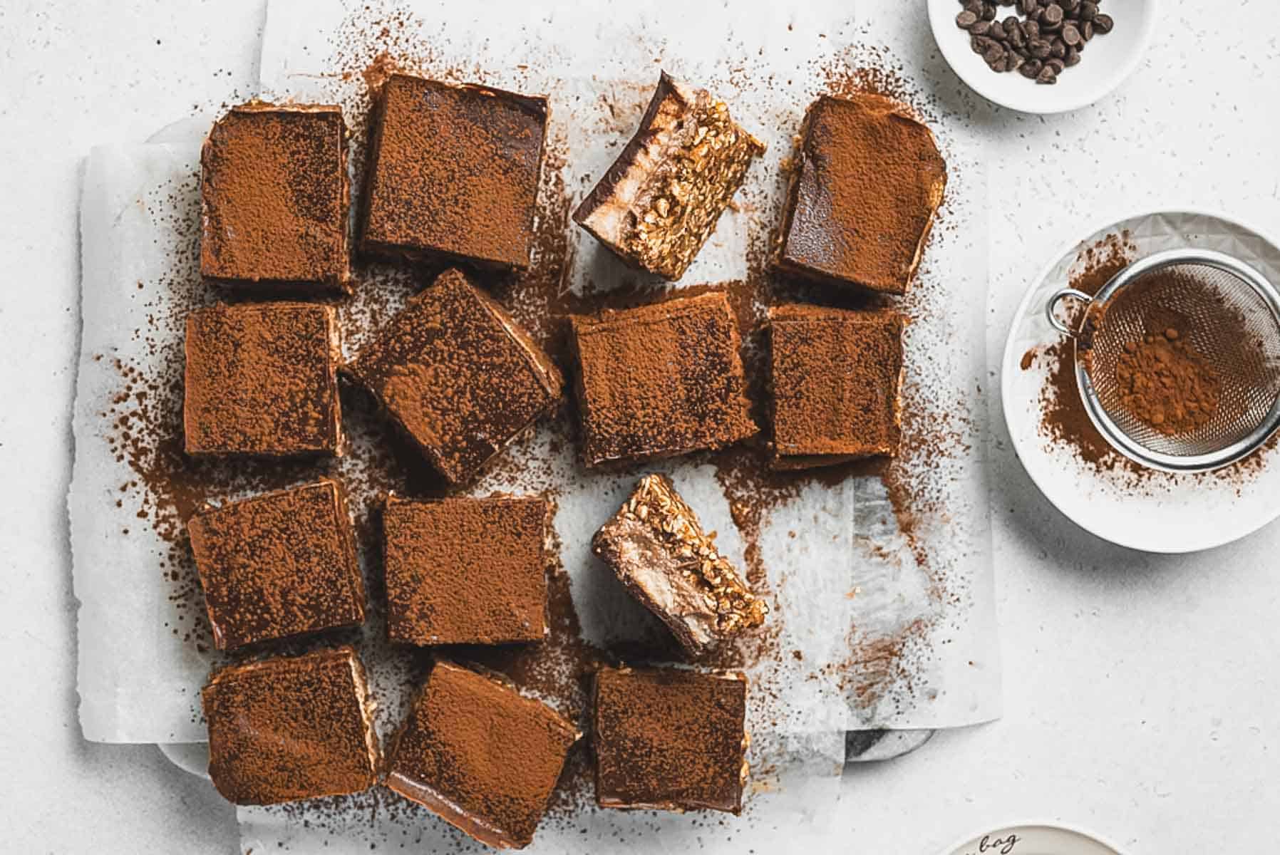 Fifteen square Tiramisu Protein Bars dusted with cocoa powder are arranged on parchment paper, with two pieces stacked to show a textured, layered interior. A sifter with cocoa and a bowl of chocolate chips are nearby.