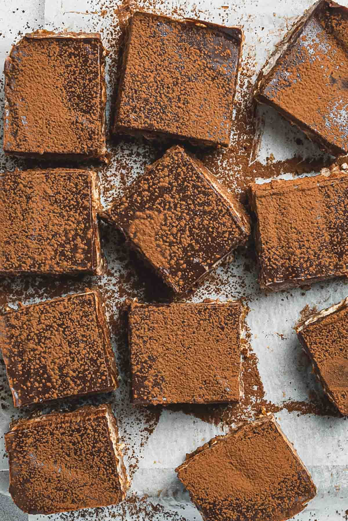 A group of Tiramisu Protein Bars, dusted with cocoa powder, are arranged on a sheet of parchment paper. The chocolate-covered dessert squares have a layered appearance and are evenly spaced.