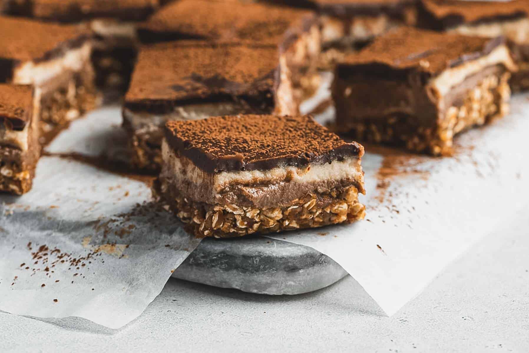 Several squares of Tiramisu Protein Bars with a base of oats, a creamy middle layer, and a chocolate topping, dusted with cocoa powder, are arranged on parchment paper atop a marble surface.